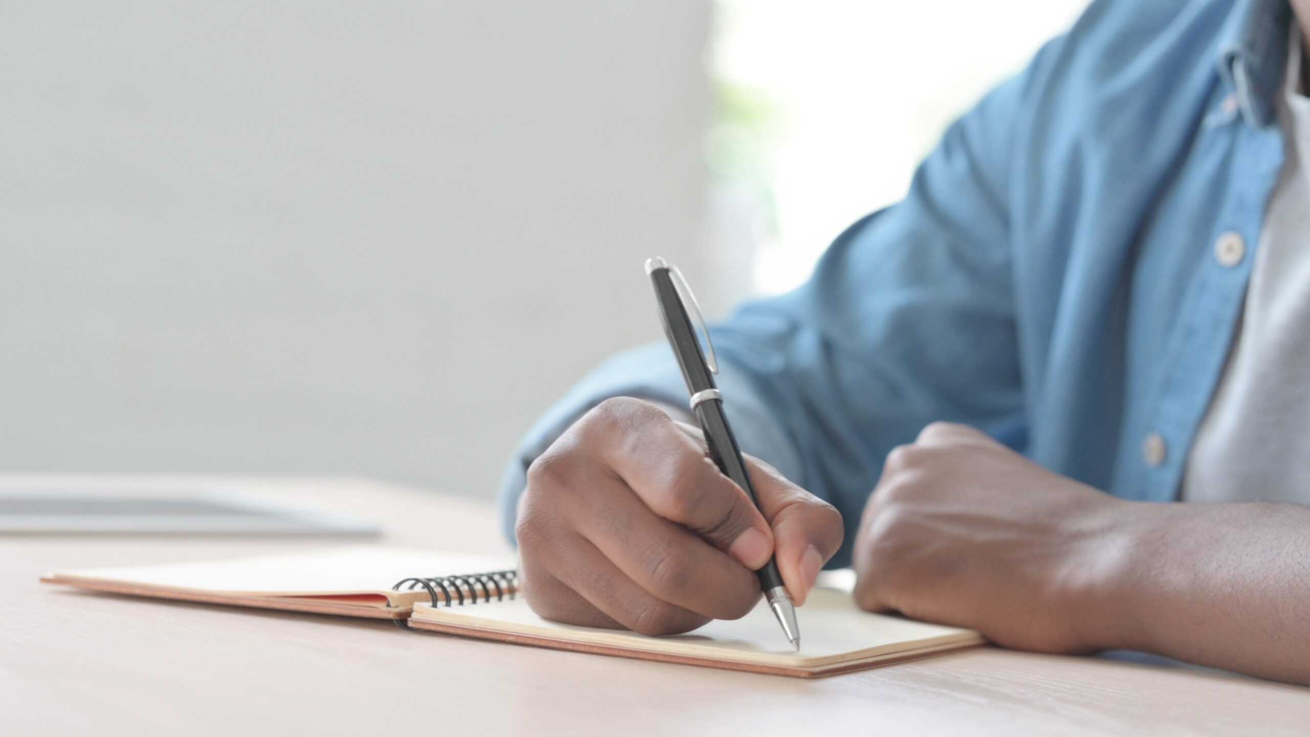 A man holding a pen, writing in a notebook while sitting at a desk.