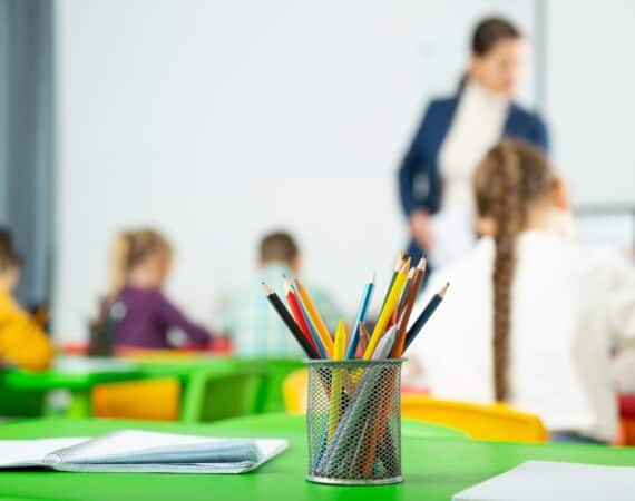 A cup of colored pencils on a table in a kindergarten classroom, while a designer is explaining UX to kids in the background.