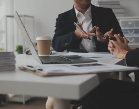 Two product managers sitting at a table with a laptop, coffee, and papers in front of them, discussing a product acquisition.