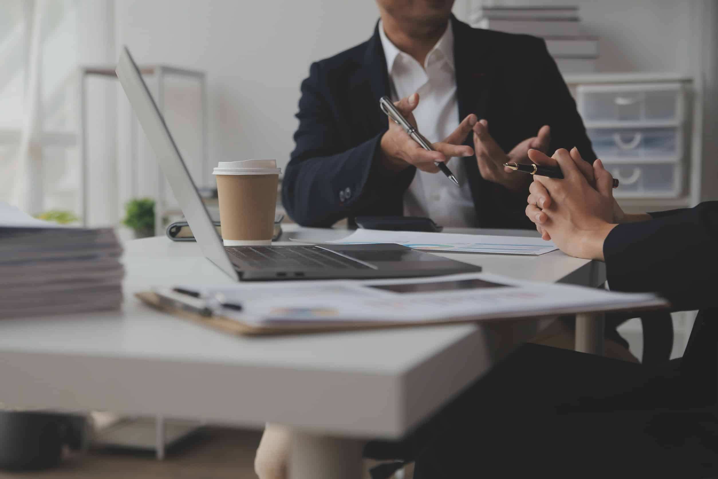 Two product managers sitting at a table with a laptop, coffee, and papers in front of them, discussing a product acquisition.