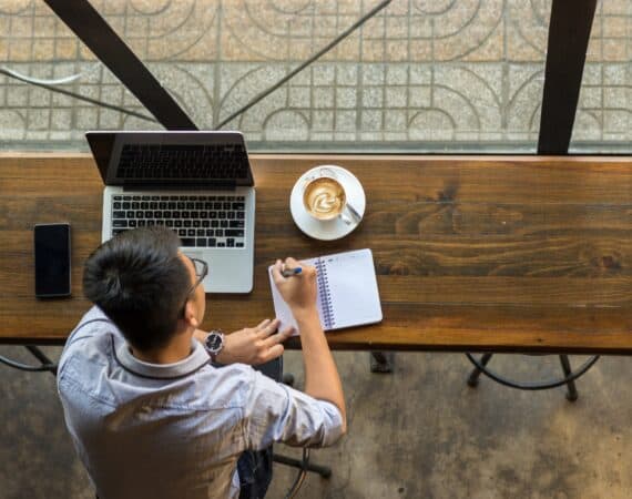 A man sitting at a window in a coffee shop, working on his laptop and writing on a notepad. This represents vulnerability to corporate counterintelligence strategies.