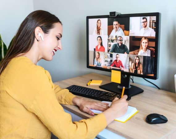 Virtual conference, morning meeting online. A young woman is using app on pc for connection with colleagues, employees, she is writing in notebook. Video call with many multiracial people togethe