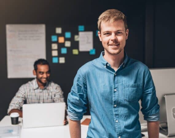 A male Product Manager in a blue shirt standing in front of a desk, while another male Product Owner sits behind the desk with a wall of sticky notes behind him.