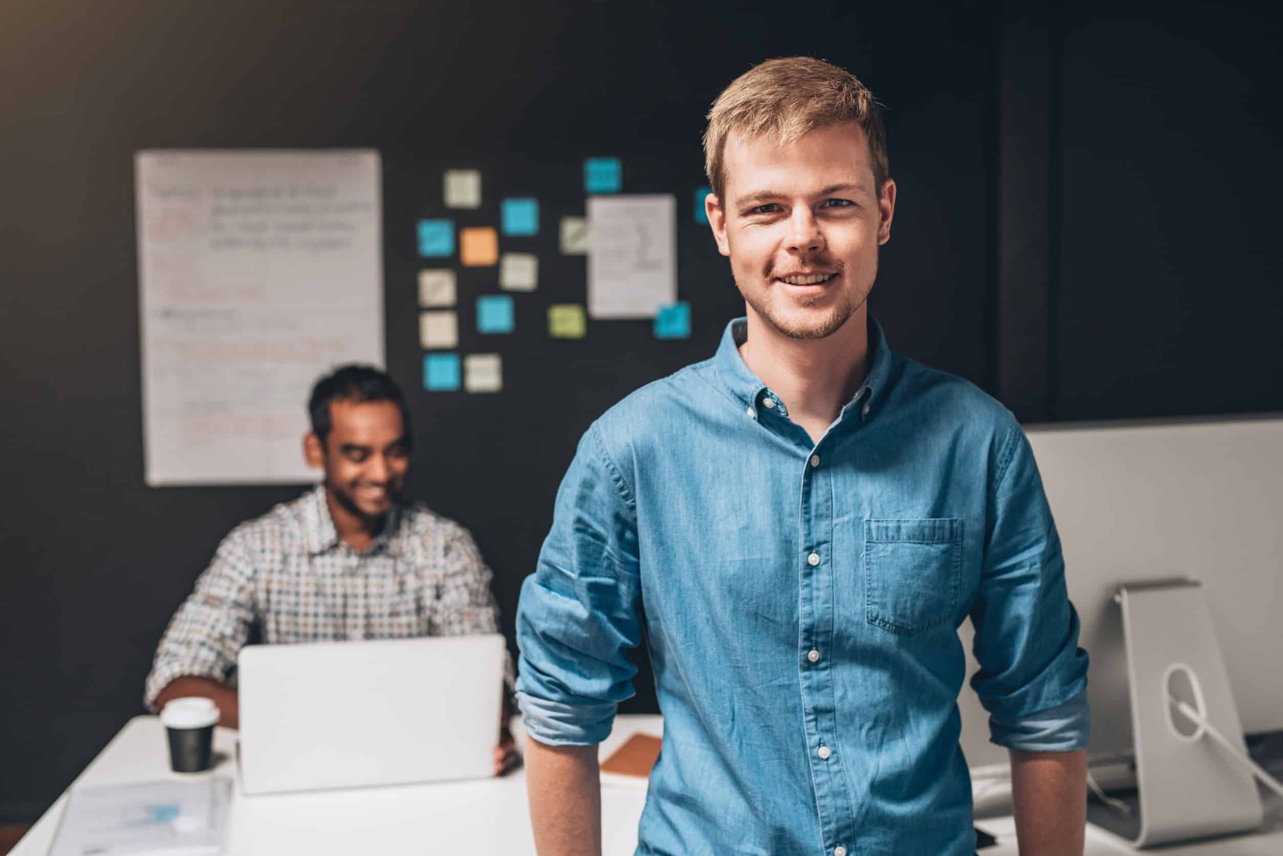 A male Product Manager in a blue shirt standing in front of a desk, while another male Product Owner sits behind the desk with a wall of sticky notes behind him.