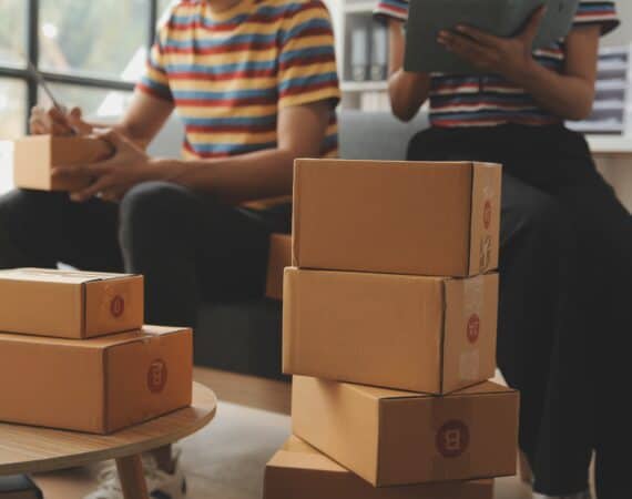 A stack of packaged boxes on a coffee table. In the background, two people sit on a couch looking at phones and tablets.