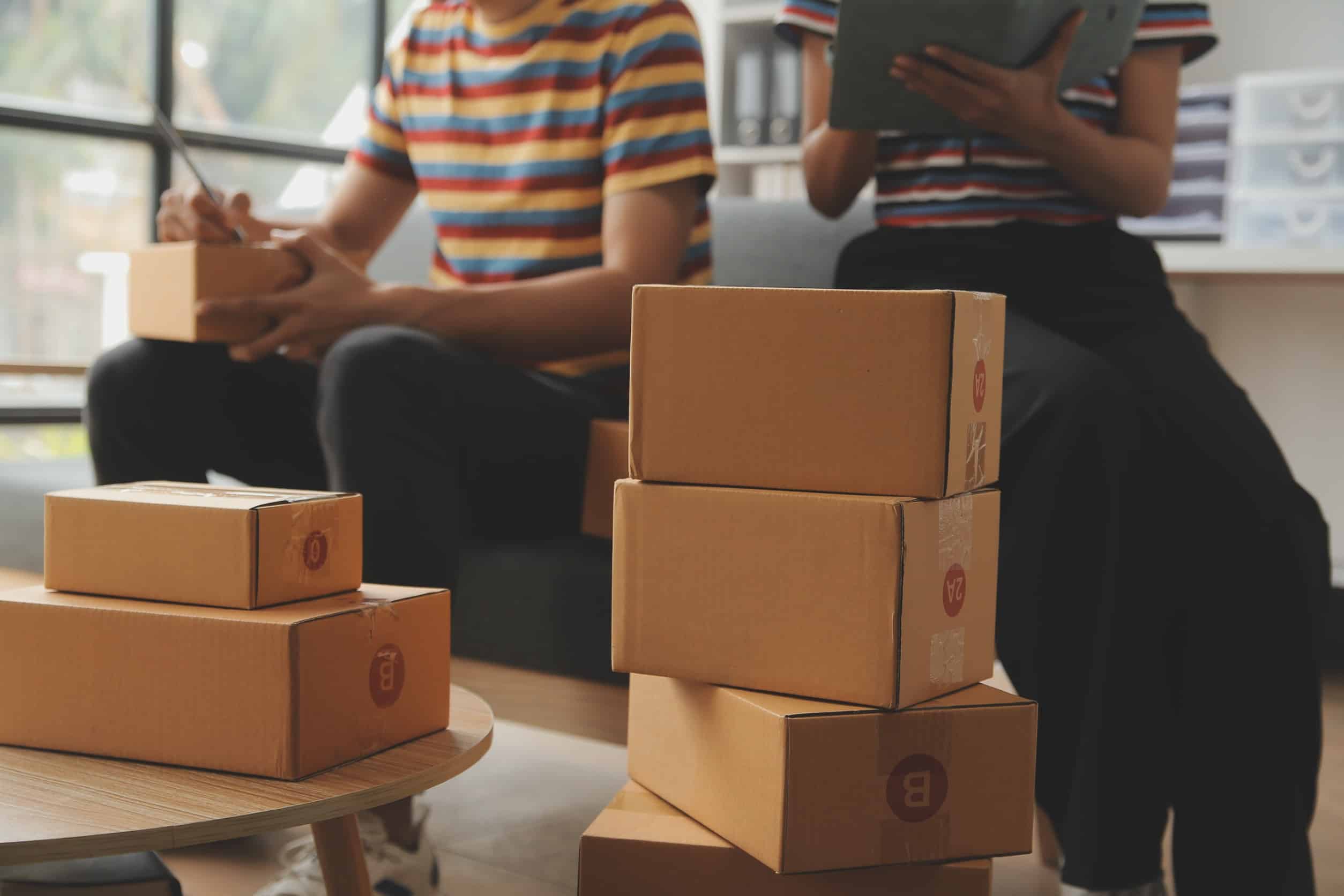 A stack of packaged boxes on a coffee table. In the background, two people sit on a couch looking at phones and tablets.