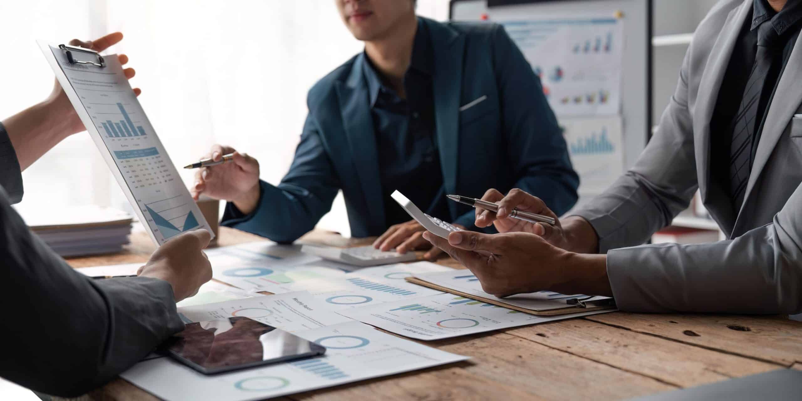 A group of business people sitting around a table with laptops and printed data reports.